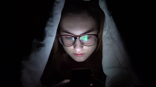 Una mujer joven yace debajo de una manta con un teléfono en sus manos. Una mujer en la oscuridad usa una almohadilla. Tableta electrónica moderna. Primer plano — Foto de Stock