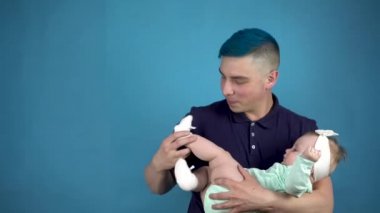Mavi saçlı genç bir baba kucağında bir bebek tutuyor. Alternatif bir adam mavi arka planda bir çocuğa çorap giydirir..