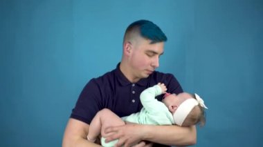 Mavi saçlı genç bir baba kucağında bir bebek tutuyor. Alternatif kişi mavi arka planda bir çocuk beşiği yapar.
