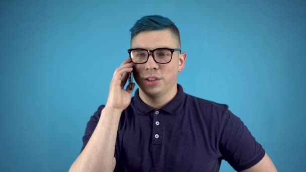 Um jovem de óculos de cabelo azul está a falar ao telefone. Pessoas alternativas com um telefone em um fundo azul. — Vídeo de Stock