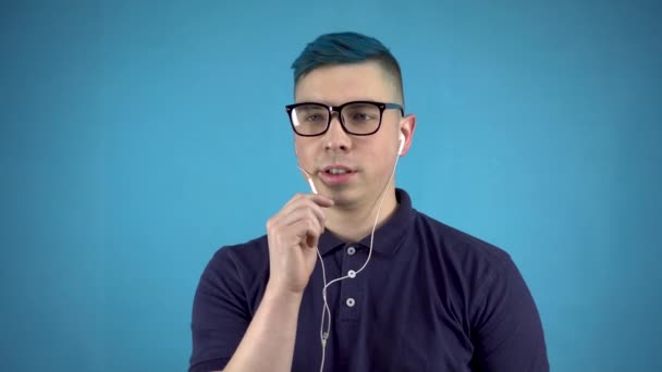 Un joven con gafas de pelo azul habla por teléfono a través de auriculares. Hombre alternativo con auriculares en orejas sobre fondo azul. — Vídeos de Stock