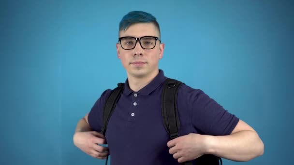 Egy kék hajú szemüveges diák hüvelykujját mutatja. Alternatív férfi egy aktatáskával a háta mögött, kék alapon. — Stock videók