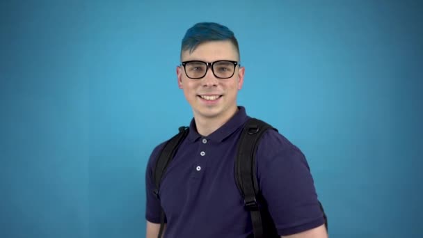 Ein Student mit Brille und blauem Haar zeigt einen Daumen. Alternativer Mann mit Aktentasche auf dem Rücken vor blauem Hintergrund. — Stockvideo