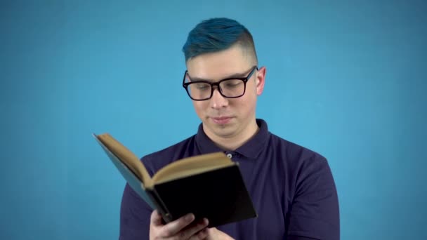 Un joven con gafas de pelo azul está leyendo un libro. Un hombre alternativo con un libro en las manos sobre un fondo azul. — Vídeos de Stock