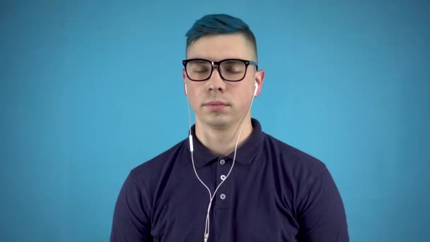 Un joven con gafas de pelo azul está escuchando música en los auriculares. Hombre alternativo con auriculares en orejas sobre fondo azul. — Vídeos de Stock