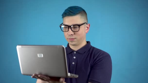 Joven con gafas de pelo azul y un portátil en las manos. Una persona alternativa está escribiendo en un netbook sobre un fondo azul. — Vídeos de Stock