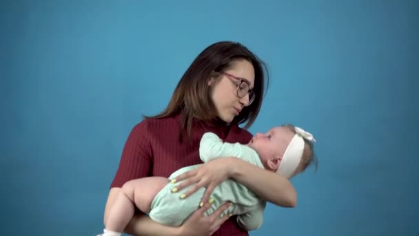 En ung mor håller ett barn i sina armar. Kvinna med en dotter på en blå bakgrund. — Stockvideo