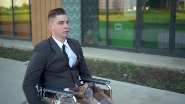 Młody biznesmen w garniturze na wózku inwalidzkim. Poważny mężczyzna jeździ na wózku inwalidzkim i patrzy na zegarek przeciwko centrum biznesowemu.. — Wideo stockowe