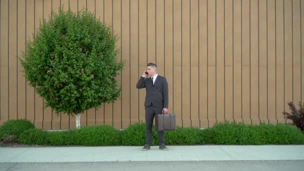 Een jonge zakenman in een pak spreekt aan de telefoon. Serieuze man met aktetas in de hand op de achtergrond van het gebouw met een bloembed. — Stockvideo