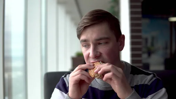 Un joven está comiendo una hamburguesa. Un hombre se sienta en un café junto a la ventana panorámica y come comida rápida . — Foto de Stock