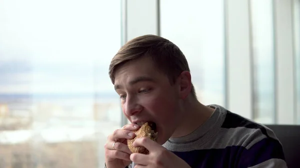Um jovem está a comer um hambúrguer. Um homem senta-se em um café pela janela panorâmica e come fast food . — Fotografia de Stock