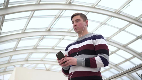 Um jovem está de pé em fones de ouvido. Um homem ouve música em fones de ouvido contra um telhado de vidro . — Fotografia de Stock