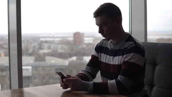 Молодой человек с телефоном сидит за столом. Мужчина сидит в кафе у панорамного окна со смартфоном в руках. Вид из окна . — стоковое фото