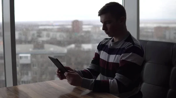 Een jongeman met een tablet zit aan een tafel. Een man zit in een café bij het panoramische raam met een tablet in zijn handen. Zicht vanuit het raam. — Stockfoto