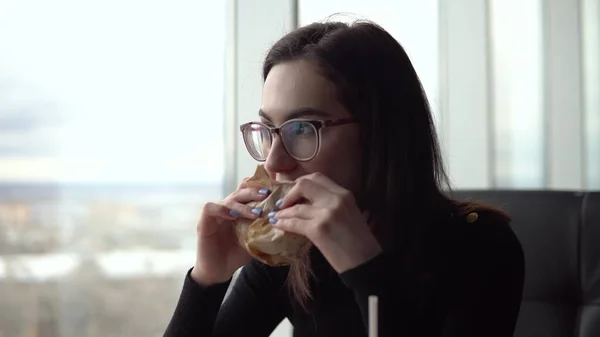 Une jeune femme mange un hamburger. Une fille s'assoit dans un café près de la fenêtre panoramique et mange de la restauration rapide . — Photo