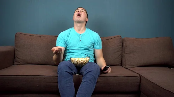 Un joven está viendo televisión y comiendo palomitas de maíz. El tipo atrapa palomitas con la boca. Sentado en el sofá. La vista desde el televisor. Mirando a la cámara —  Fotos de Stock