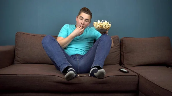 A young man is watching TV and eating popcorn. The guy is eating fallen popcorn on the sofa. Sitting on the couch. The view from the TV. Looking at the camera — ストック写真