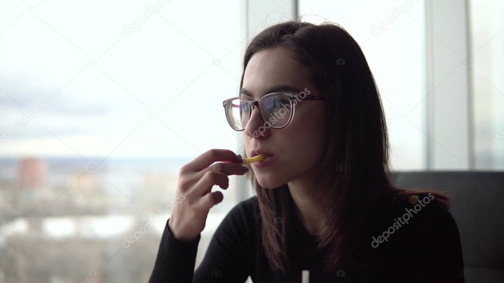 Young woman eating fried potatoes. A girl sits in a cafe by the panoramic window and eats fast food.