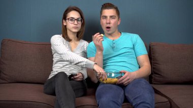 Bir erkek ve bir kadın televizyon izleyip gülüyorlar. Genç bir çift kanepede oturuyor ve cips yiyorlar. Ellerinde kırıntılar olan cam bir tabak. Televizyondaki manzara. Kameraya bakıyorum.