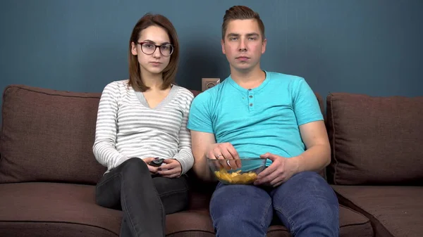 Ein Mann und eine Frau sehen fern und lachen. Ein junges Paar sitzt auf der Couch und isst Chips. Glasplatte mit Chips in den Händen. Der Blick aus dem Fernseher. Blick in die Kamera — Stockfoto