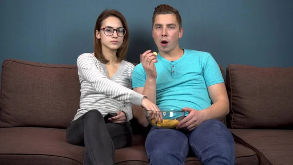 Ein Mann und eine Frau sehen fern und lachen. Ein junges Paar sitzt auf der Couch und isst Chips. Glasplatte mit Chips in den Händen. Der Blick aus dem Fernseher. Blick in die Kamera — Stockfoto