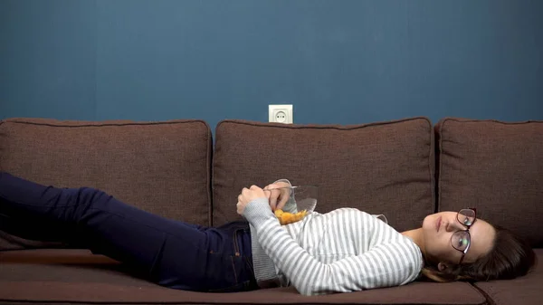 Una joven con gafas come papas fritas y ve televisión. Chica acostada en el sofá — Foto de Stock