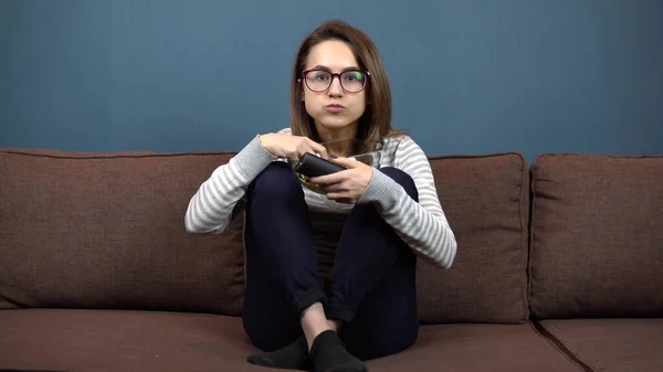 Una joven con gafas come papas fritas y ve televisión. Chica sentada con las piernas en el sofá — Foto de Stock