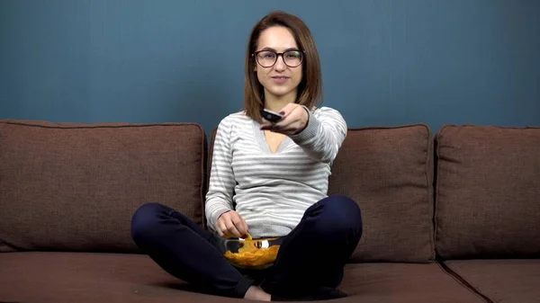 Una joven con gafas come papas fritas y ve televisión. Chica sentada con las piernas en el sofá —  Fotos de Stock