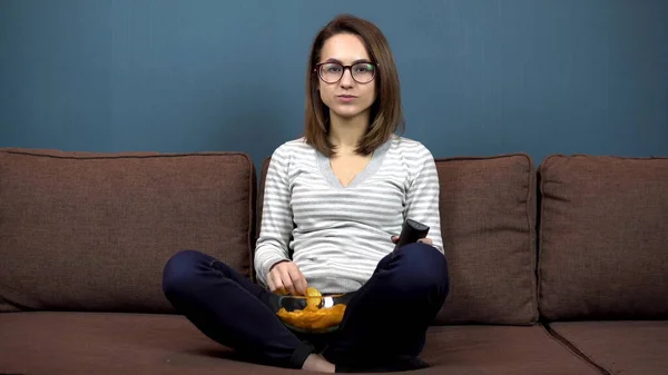 Una joven con gafas come papas fritas y ve televisión. Chica sentada con las piernas en el sofá —  Fotos de Stock