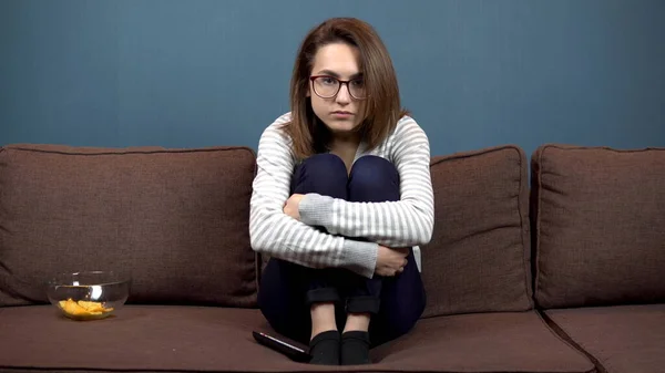 Una joven con gafas lee nerviosamente viendo la televisión. La chica se subió al sofá y abrazó sus piernas. Nervioso al ver la televisión —  Fotos de Stock