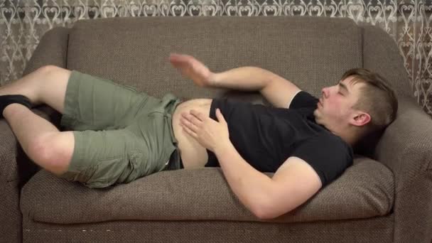 Ein junger Mann liegt auf einem Sofa und klopft sich auf den Bauch. Übergewicht in Quarantäne. — Stockvideo
