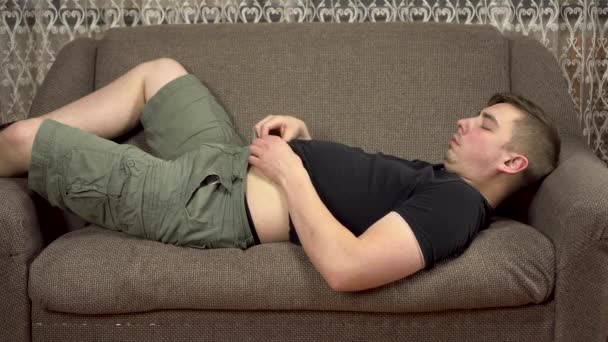 Молодий чоловік лежить на дивані, торкаючись його товстого живота. Надмірна вага в карантині . — стокове відео