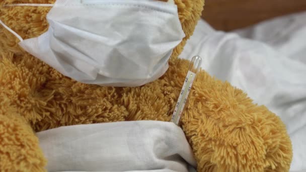 A teddy bear measures the temperature with a mercury thermometer. The doctor takes the thermometer closeup. The bear lies in bed with a medical mask. — Stock Video