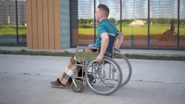 Un joven en silla de ruedas. Un hombre cabalga en una silla de ruedas sobre el fondo de un edificio de cristal. Transporte especial para minusválidos . — Vídeos de Stock