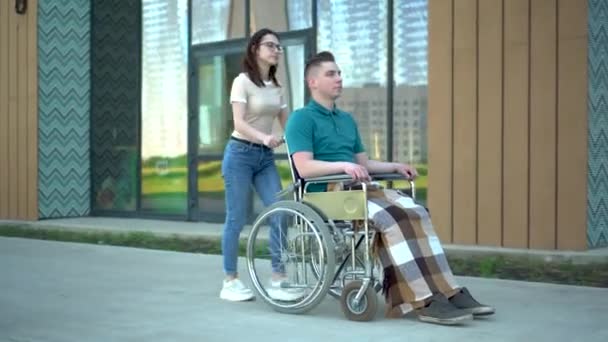 Una joven lleva a un joven en silla de ruedas. Un asistente lleva a una persona con discapacidad en una silla de ruedas a lo largo de la calle. Transporte especial para minusválidos . — Vídeos de Stock