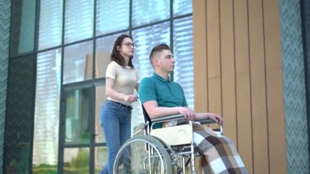 Een jonge vrouw draagt een jonge man in een rolstoel. Een assistent draagt een gehandicapte in een rolstoel langs de straat. Speciaal vervoer voor gehandicapten. — Stockvideo