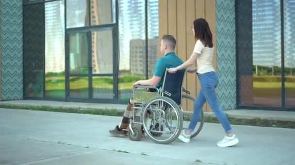 若い女性が若い男を車椅子に乗せている。アシスタントは路上で車椅子の障害者を運んでいる。障害者のための特別輸送. — ストック動画