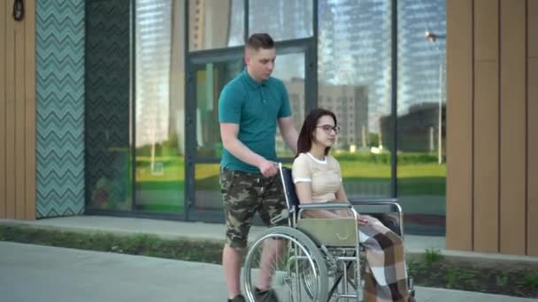 Молодий чоловік носить молоду жінку в інвалідному візку. Асистент несе інваліда в інвалідному візку вздовж вулиці. Спеціальний транспорт для інвалідів . — стокове відео