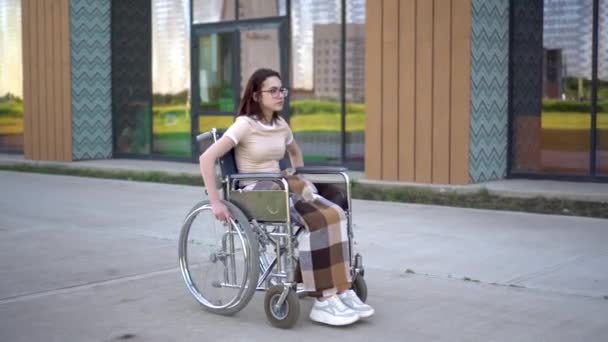 若い女性が車椅子に乗って心臓発作が起こります。ガラスの建物を背景に車椅子の少女が心をつかむ。障害者のための特別輸送. — ストック動画