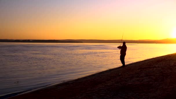Bir adam gün doğumunda nehir kenarında balık tutuyor. Sarı güneş ufuktan yükseliyor. Bir balıkçının silueti. Adamın biri olta atıyor.. — Stok video