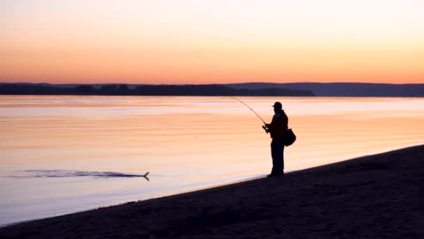 Egy ember fogott egy halat. Egy férfi sziluettje, aki napkeltekor horgászik a folyónál. A sárga nap felkel a horizontból.. — Stock videók
