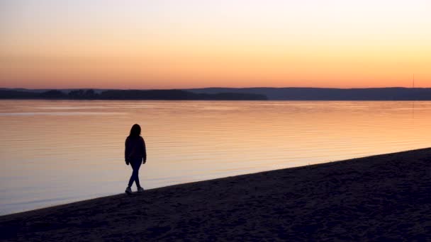 Egy fiatal nő sétál a parton a folyó mentén napkeltekor. Egy lány sziluettje. A hajnal felé tart.. — Stock videók