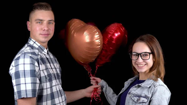 Ein junger Mann überreicht einer jungen Frau herzförmige Luftballons. Mann und Frau küssten sich auf schwarzem Grund. Valentinstag ist der Tag aller Verliebten. — Stockfoto