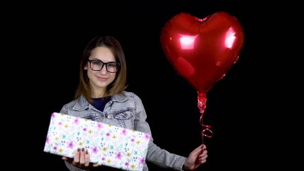 A young girl is standing with a gift and heart-shaped balloons. A woman holds in her hands a gift and balls with helium on a black background. Valentines Day is the day of lovers.