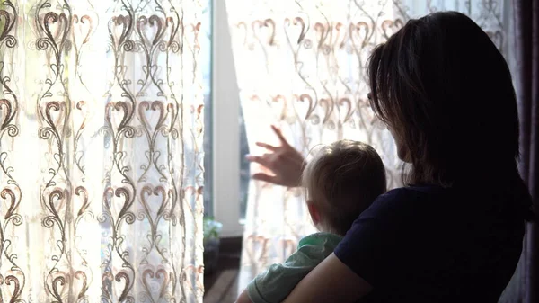 Mladá matka drží dítě v náručí. Žena s dítětem, co se dívá z okna a zatahuje závěs.. — Stock fotografie