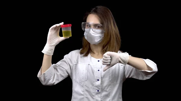 La jeune femme médecin vérifie les analyses d'urine. La femme examine le bocal de test, puis montre un pouce vers le bas. Analyse d'urine négative. Sur fond noir. — Photo