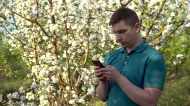 Doğada elinde telefon olan genç bir adam. Bir adam, çiçek açan bir ağacın arka planında duran bir akıllı telefondan çalışıyor..