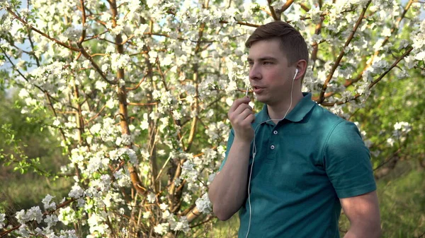 Joven en la naturaleza con auriculares. Un hombre está hablando por teléfono a través de auriculares mientras está de pie contra un árbol floreciente . —  Fotos de Stock