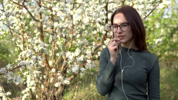 Mujer joven en la naturaleza con auriculares. Una chica está hablando por teléfono a través de auriculares mientras está de pie contra un árbol floreciente . —  Fotos de Stock