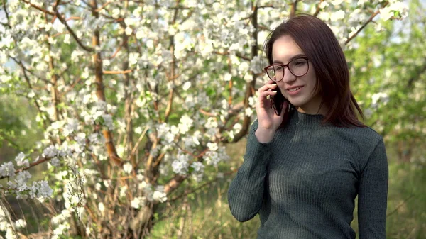 Jovem mulher na natureza com um telefone. Uma menina está falando ao telefone enquanto está de pé contra uma árvore florida . — Fotografia de Stock